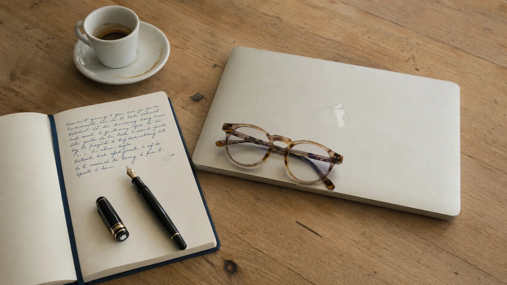 Tortoiseshell blue light glasses resting on closed MacBook Air with Montblanc Meisterstück fountain pen, notebook and espresso on natural oak desk